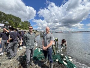 Two adult men, members of Seaspan's Environment team, stand beside each other smiling. They are wearing rain boots and are standing on a rocky beach adjacent to the Burrard Inlet and MacKay Creek as part of an educational opportunity at MacKay Creek organized by the Coho society. There is a large group of students behind them and some volunteers with a seine net standing inside the water and collecting marine samples.