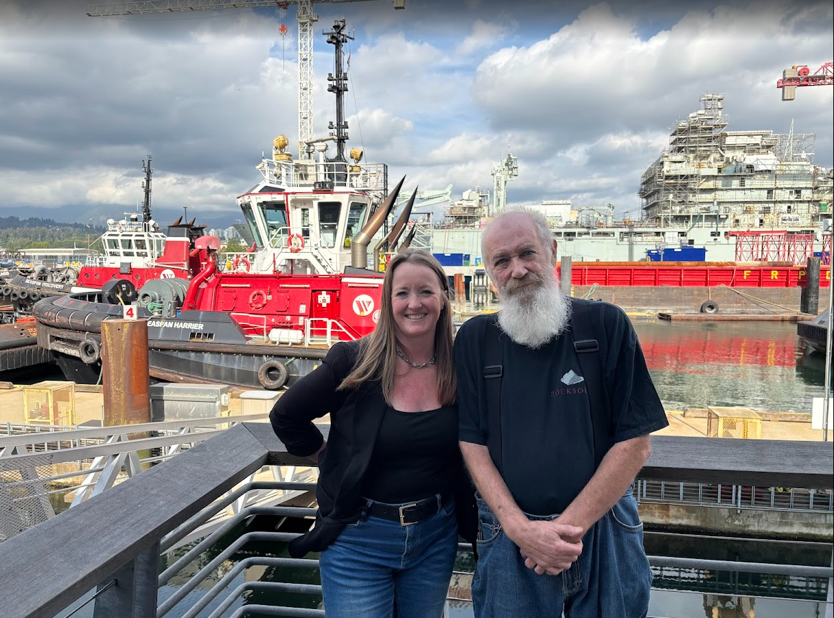 Two people standing outside on a dock, in front of a tugboat
