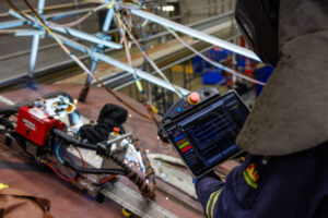 A Seaspan welder holding a tablet, robotic welding robot in background.