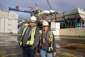 Two employees wearing full PPE and white hard hats stand in front of the second joint support ship under construction at Vancouver Shipyards. On the left is Richard Thomasson, and on the right is Dalyce Temple. Both are smiling. 