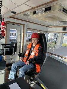 Young boy in hard hat and life vest sits in tugboat captain chair