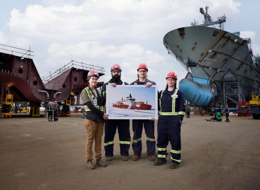 Four shipyard employees wearing PPE hold a large photo of an icebreaker. They are in a shipyard with a large ship, JSS2, and a ship block behind them
