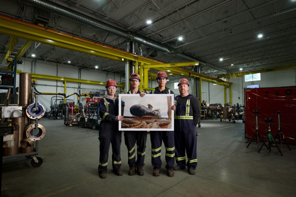 Four shipyard employees in full PPE (hardhats, coveralls, safety glasses) hold a large photo of a submarine with an industrial warehouse background