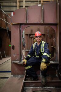 A ship fitter with a hard hat and a high vis vest sits on a ship block